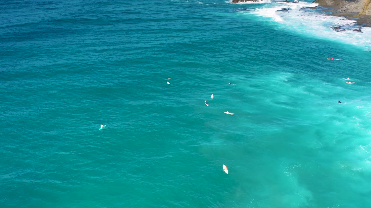 Wide aerial frame over Cerritos, Baja California Sur, with the rocky wave line at the upper right, clear blue water filling the frame, tiny boards offshore, and no visible people.