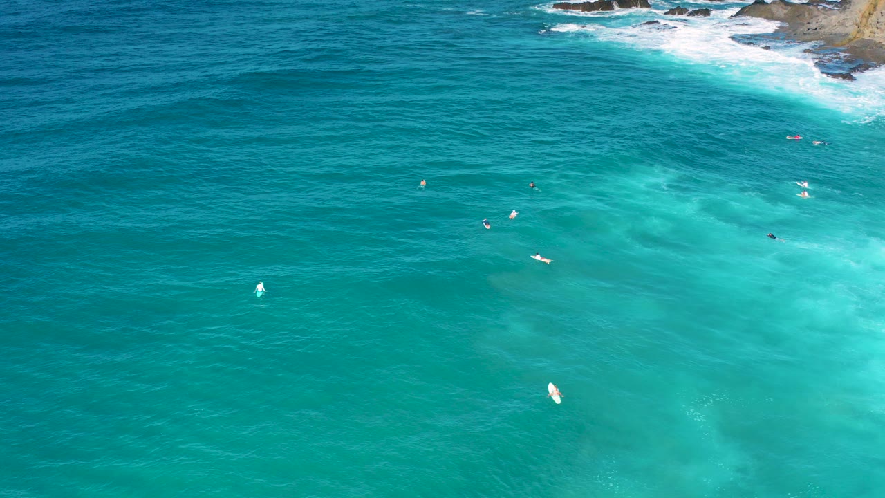 Aerial frame over Cerritos, Baja California Sur showing turquoise water, faint underwater sand color, tiny surfers or boards, rocky shoreline at the top edge, and no visible faces.