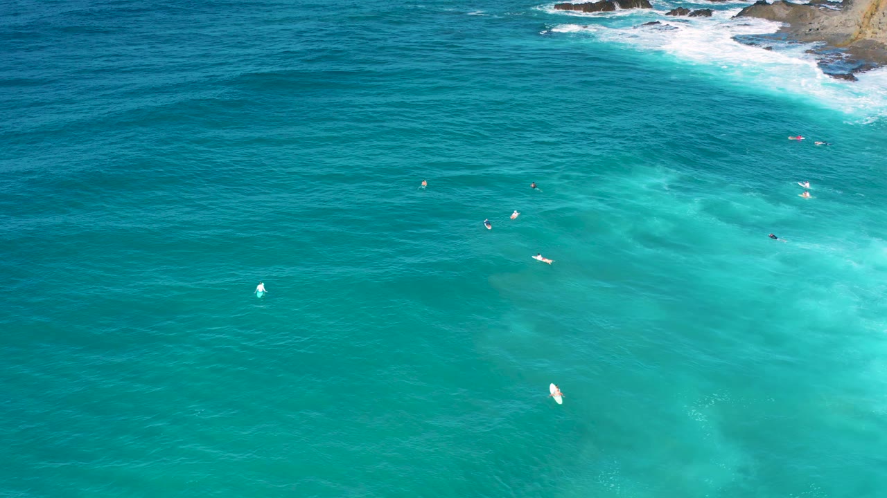 Aerial view over Cerritos water where blue-green bands fill the frame, a few tiny surfers or boards sit in the midground, and white wash touches the rocky shoreline near the top.