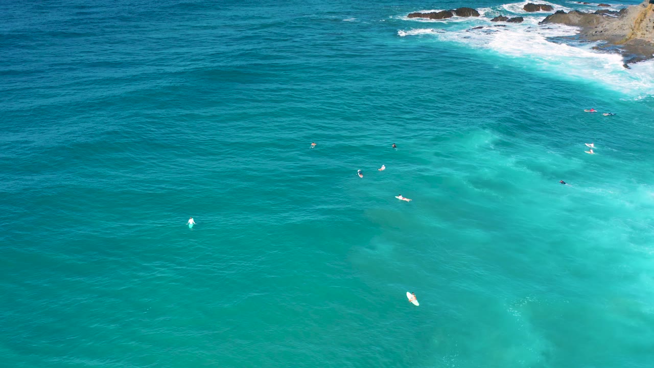 The frame holds a high drone-like view of Cerritos ocean, with turquoise shallows on the right, darker blue water on the left, scattered tiny boards, and rocky surf at the top.