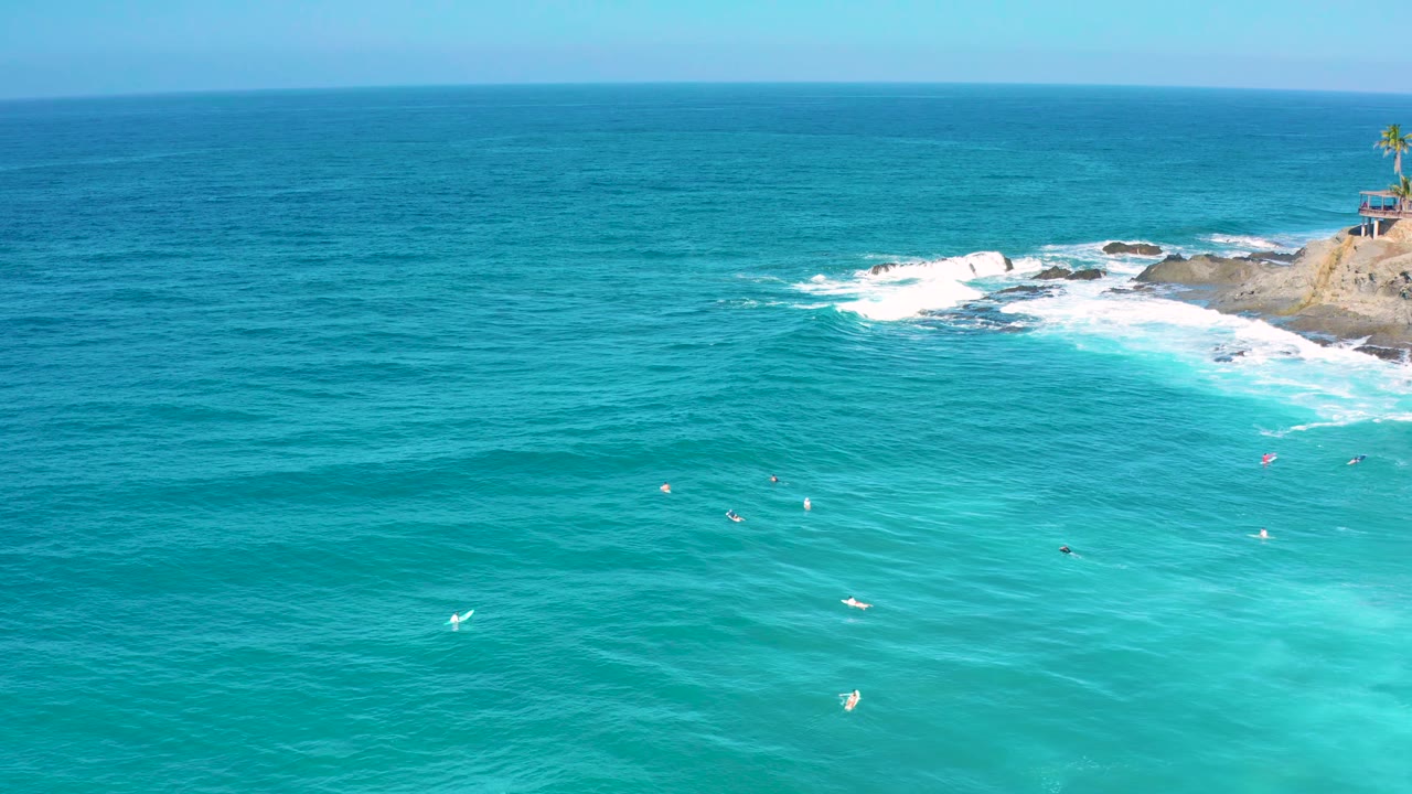 Aerial frame from Cerritos, Baja California Sur showing rippled blue water, small white wave patches, and the rocky point with a palm-topped structure near the upper right, with no visible people.