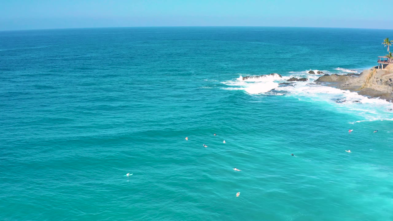 High drone view over teal water in Cerritos, Baja California Sur, with scattered whitecaps and surf hitting the rocky shore at the upper right, with no visible people.