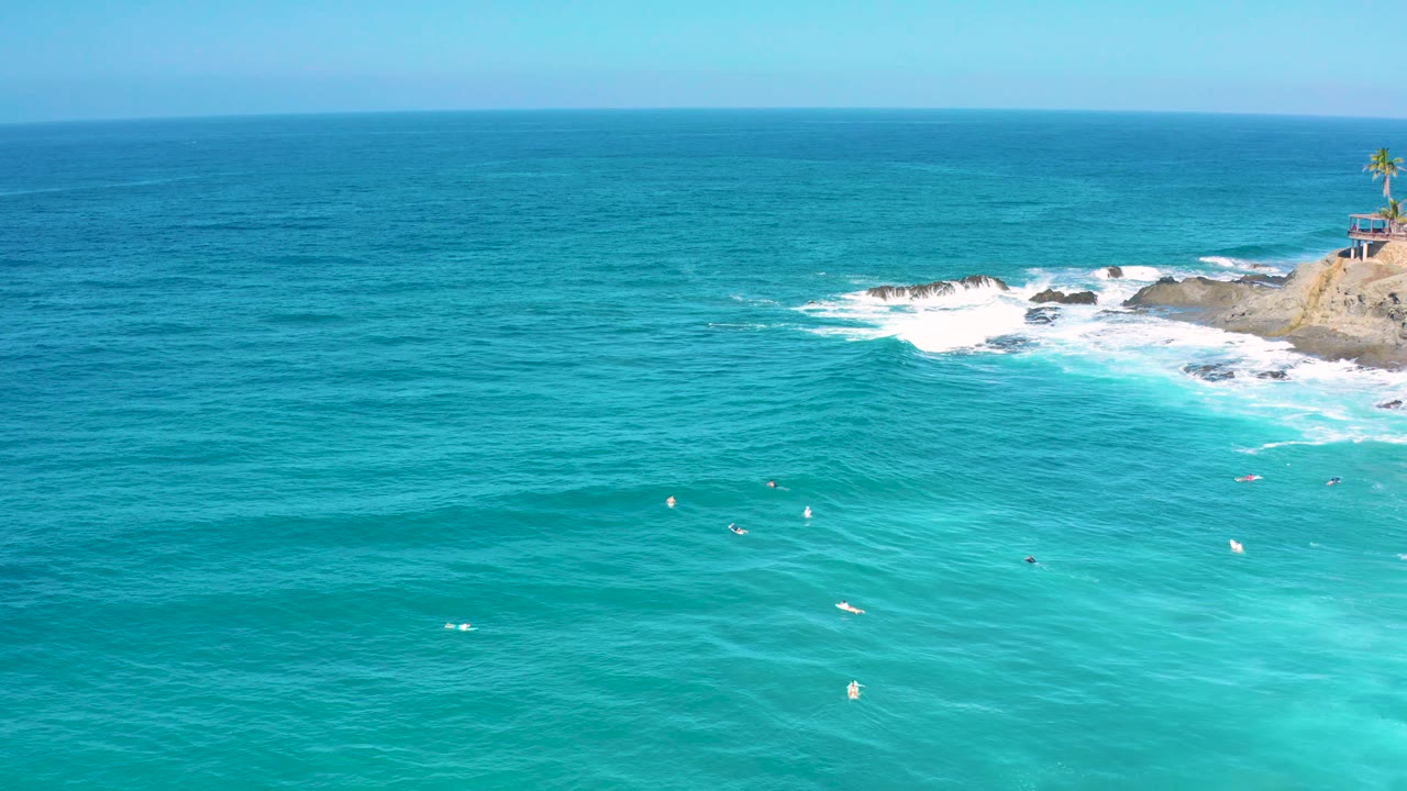 Bright aerial view in Cerritos, Baja California Sur with turquoise foreground water, white surf wrapping the rocks, and a small palm-topped lookout above the shore, with no visible people.