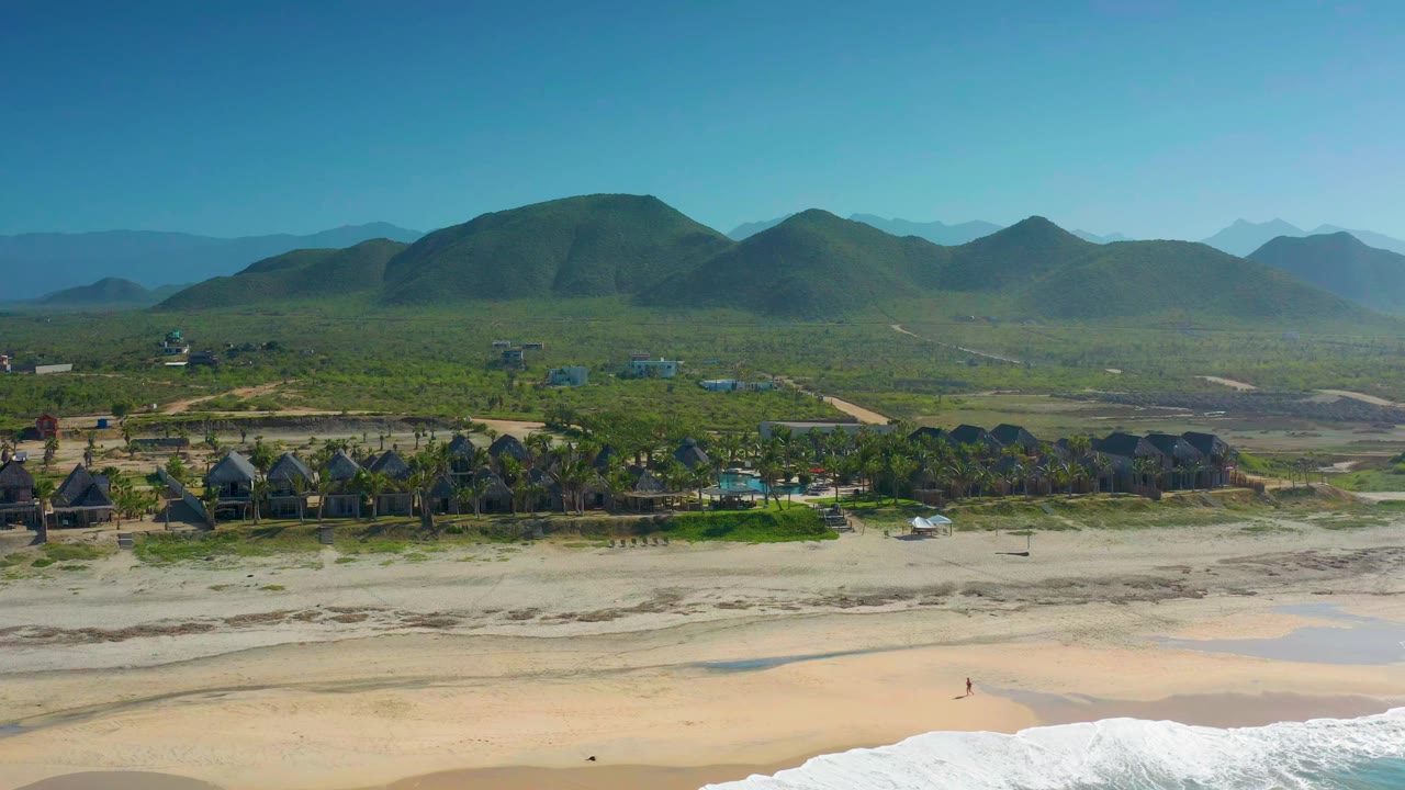 Opening aerial frame over Cerritos, Baja California Sur, showing white surf at the bottom edge, a broad sand beach, palm-lined buildings, green hills, and no recognizable people.
