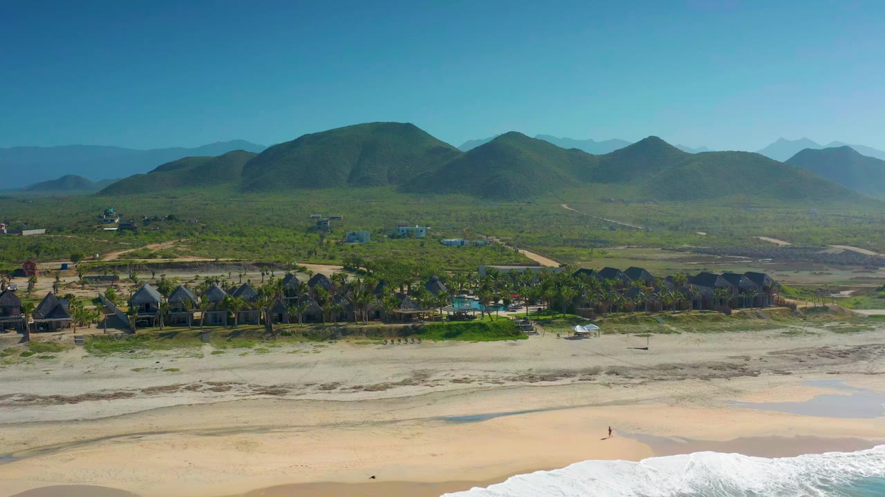Aerial frame over Cerritos with the wave line shifted along the lower edge, pale sand in front, palms and beach buildings across the middle, and hazy green mountains beyond.