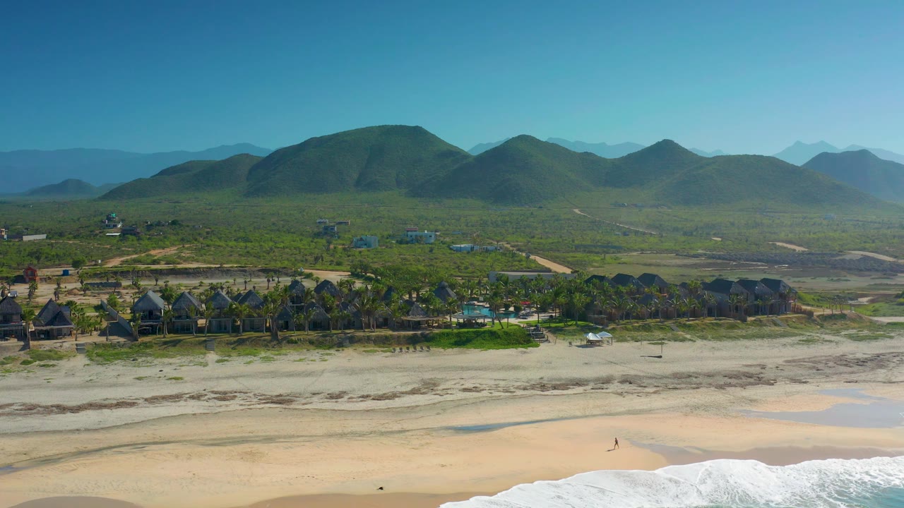 Cerritos aerial view with foamy surf entering from the lower right, open sand across the foreground, a thin row of palms and buildings, and rounded mountains behind.