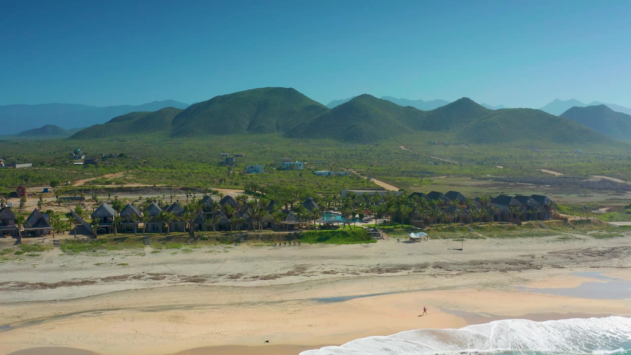 Elevated beach view with waves, sand flats, trees, low buildings, and hills.
