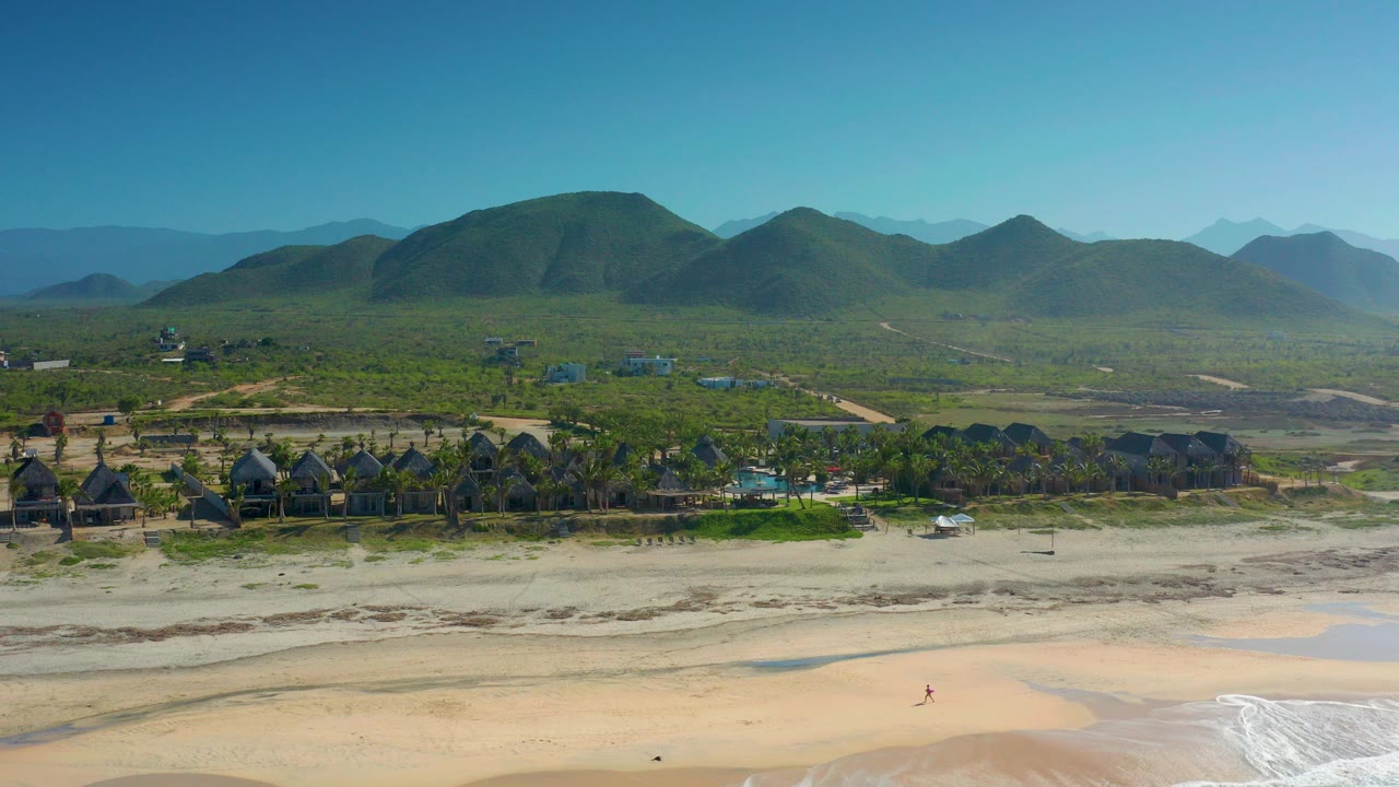 Wide aerial frame of Cerritos, Baja California Sur showing pale sand, white surf at the lower edge, hut-like buildings and palms across the shoreline, green mountains behind, and no visible people.