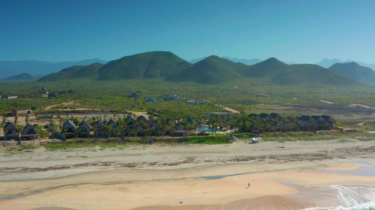 Wide Cerritos beach frame with a smooth band of sand, small white waves at the bottom right, a row of dark-roof huts among palms, hazy green mountains, and no visible people.