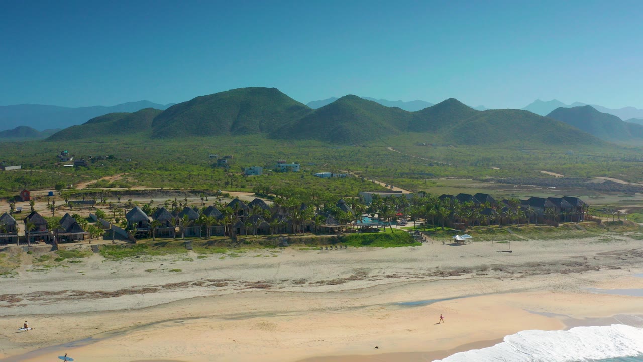 Wide aerial frame of Cerritos, Baja California Sur with pale beach and white surf below, palm-lined buildings beyond the sand, green hills and layered mountains in the distance, and no visible people.