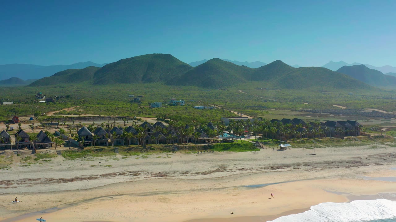 Aerial view over Cerritos, Baja California Sur showing a low wave line at the bottom edge, open sand, palm rows and dark roof structures, green hills, hazy distant ridges, and no visible people.