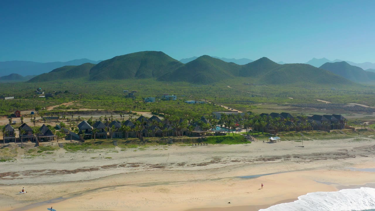 High coastal frame from Cerritos, Baja California Sur with the surf shifted toward the lower right, broad sand across the foreground, palm-lined structures in the midground, green hills, and no visible people.