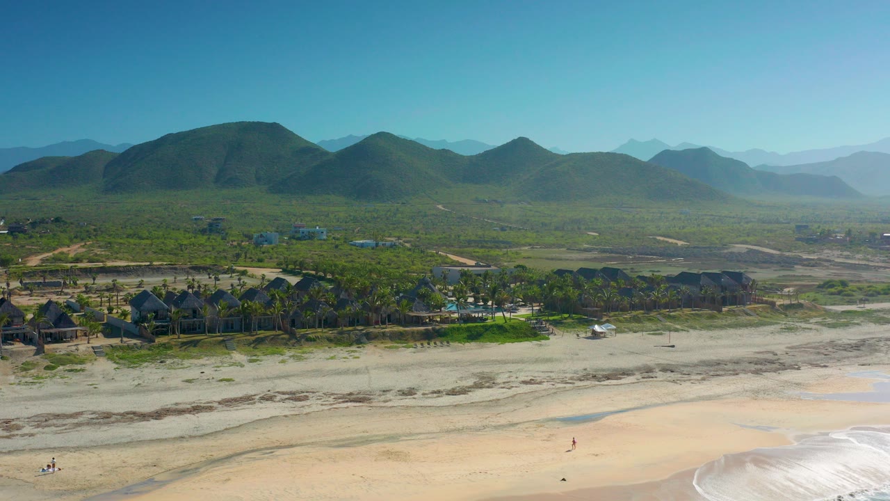 Wide aerial view in Cerritos, Baja California Sur, Mexico, showing pale beach, low surf, palm trees, small buildings, and green mountains under clear sky with no recognizable people visible.