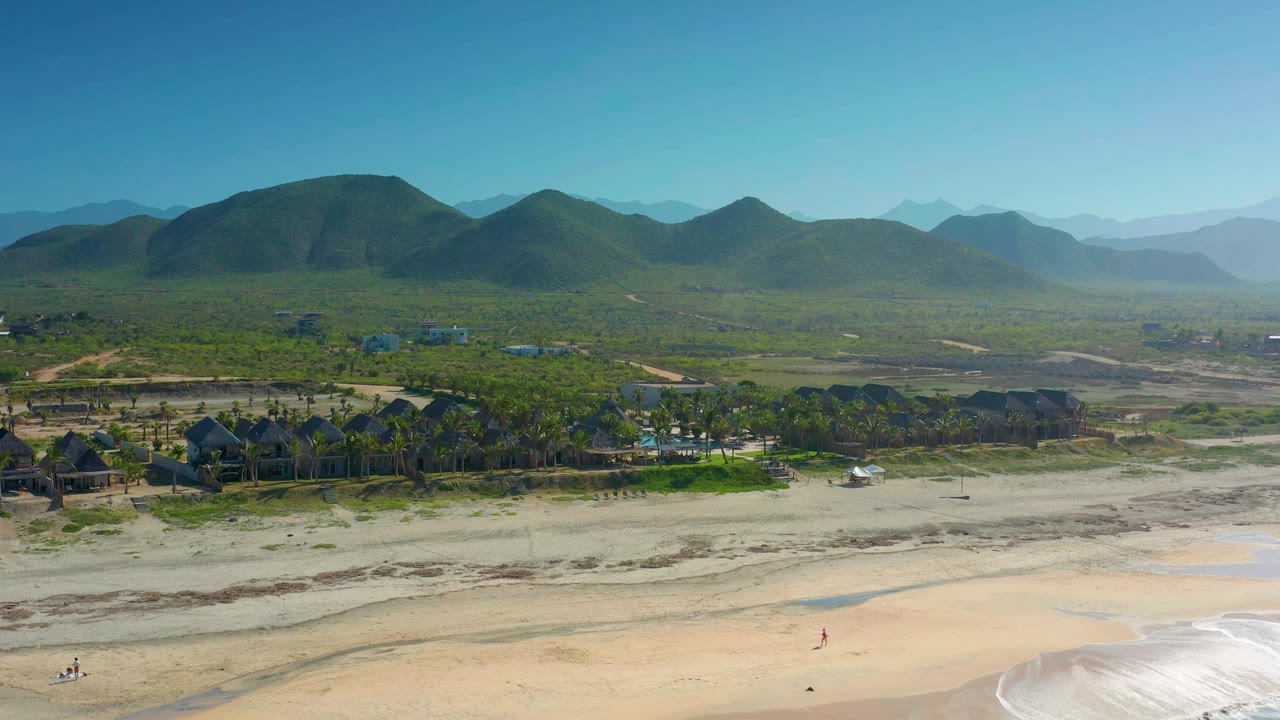Aerial frame over Cerritos beach with the palm-lined buildings slightly more centered, pale sand below, and layered mountains fading into the background haze with no recognizable people visible.