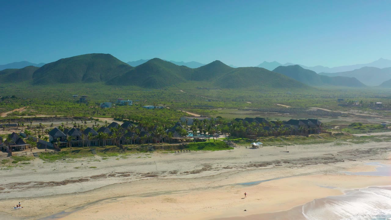 Elevated Cerritos coastline view with foamy water at the lower edge, open sand, clustered palms and roofs, and green mountains across the horizon with no recognizable people visible.