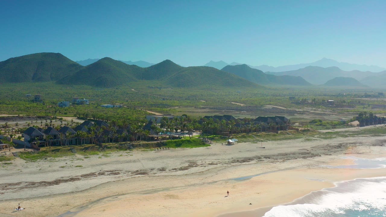 Opening frame of Cerritos, Baja California Sur with pale beach sand and surf in front, a palm-lined property in the midground, and green mountains across the horizon with no recognizable people visible.
