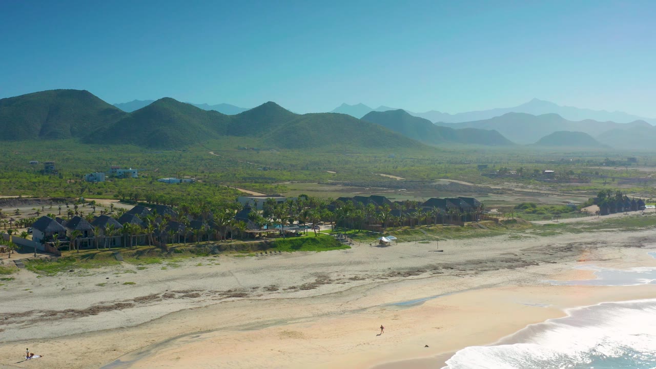 Wide Cerritos beach frame showing a pale shoreline and white water at the bottom edge, green trees and scattered structures behind the sand, and a long mountain ridge under bright blue sky.