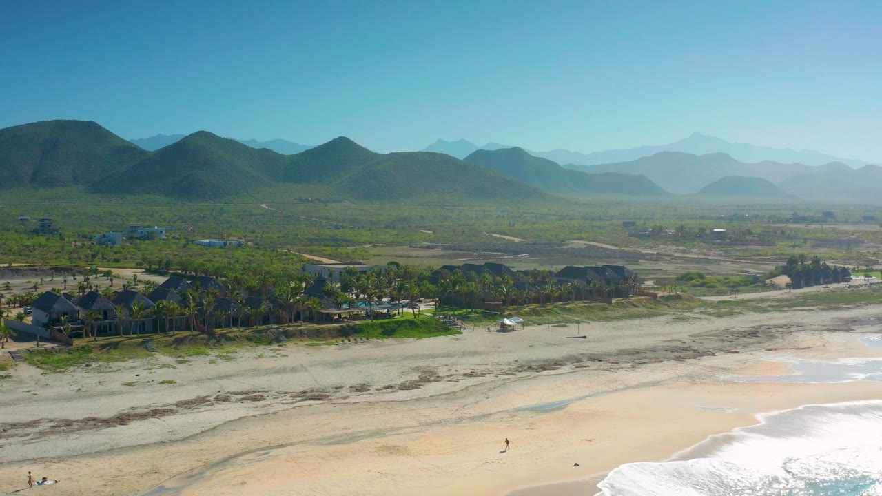 Beachfront aerial with mountains, palms, and light surf.