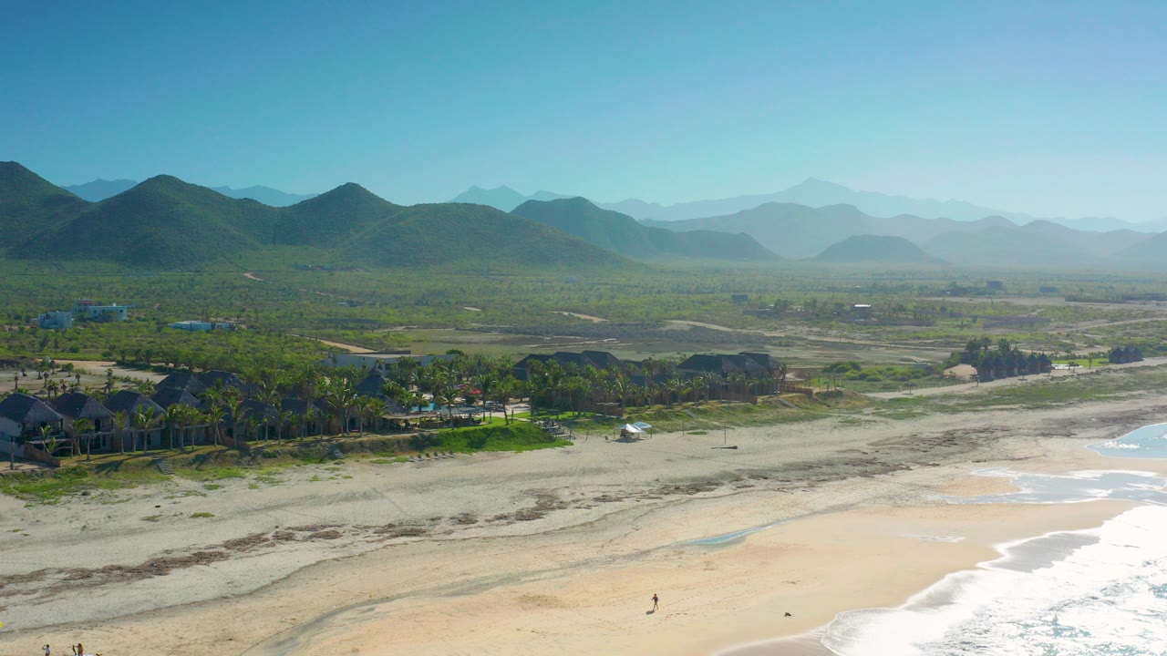 Wide aerial view over Cerritos, Baja California Sur, with white surf on pale sand, palms and low houses inland, green hills, hazy mountains, and no visible people.