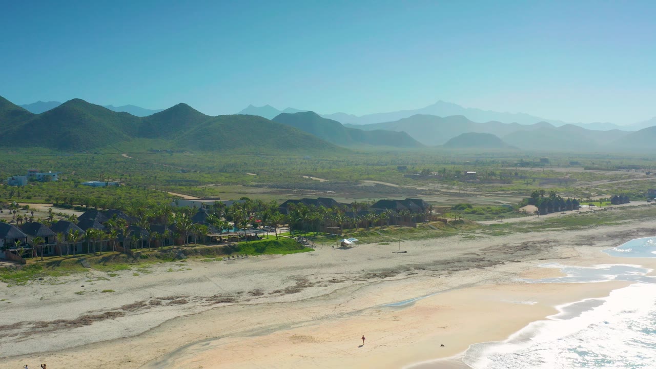 High drone view in Cerritos with whitewash crossing the lower right, broad sand, a cluster of palms and low buildings, green hills, blue haze, and no visible people.