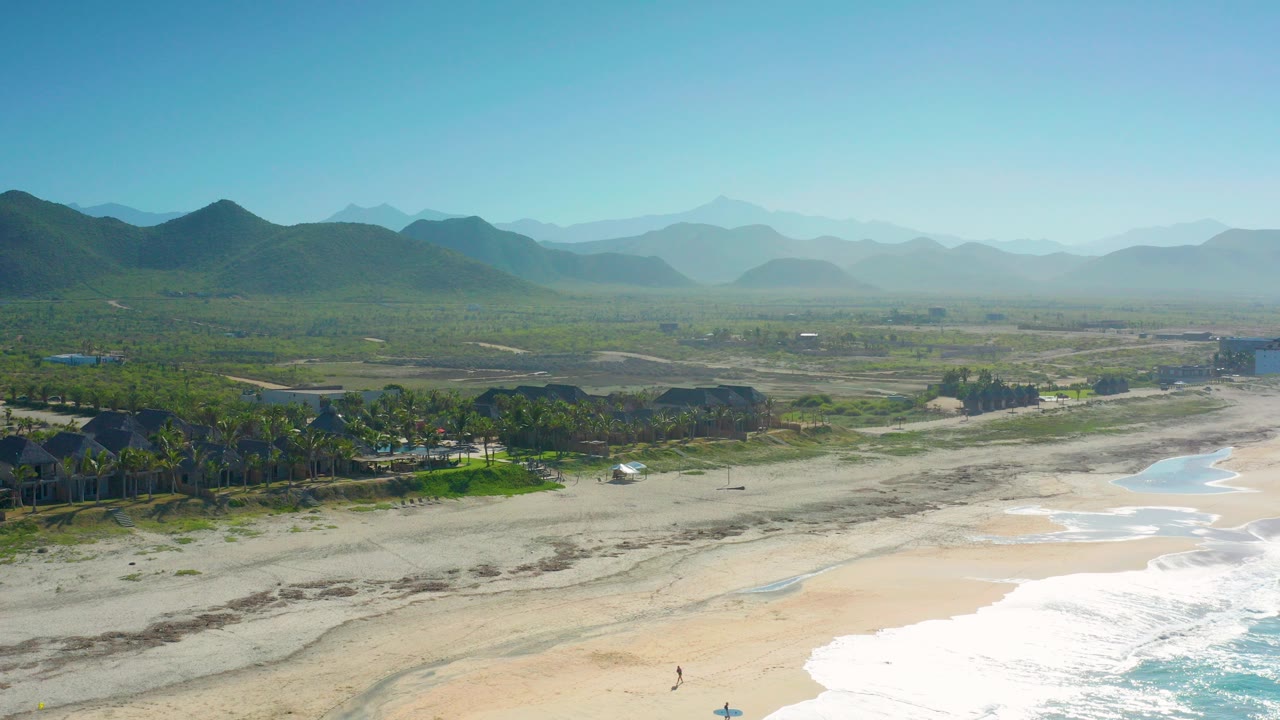 Aerial frame from Cerritos, Baja California Sur showing surf at the lower right, pale sand and a remote road across the foreground, palms and buildings in the midground, hazy hills behind, and no visible people.