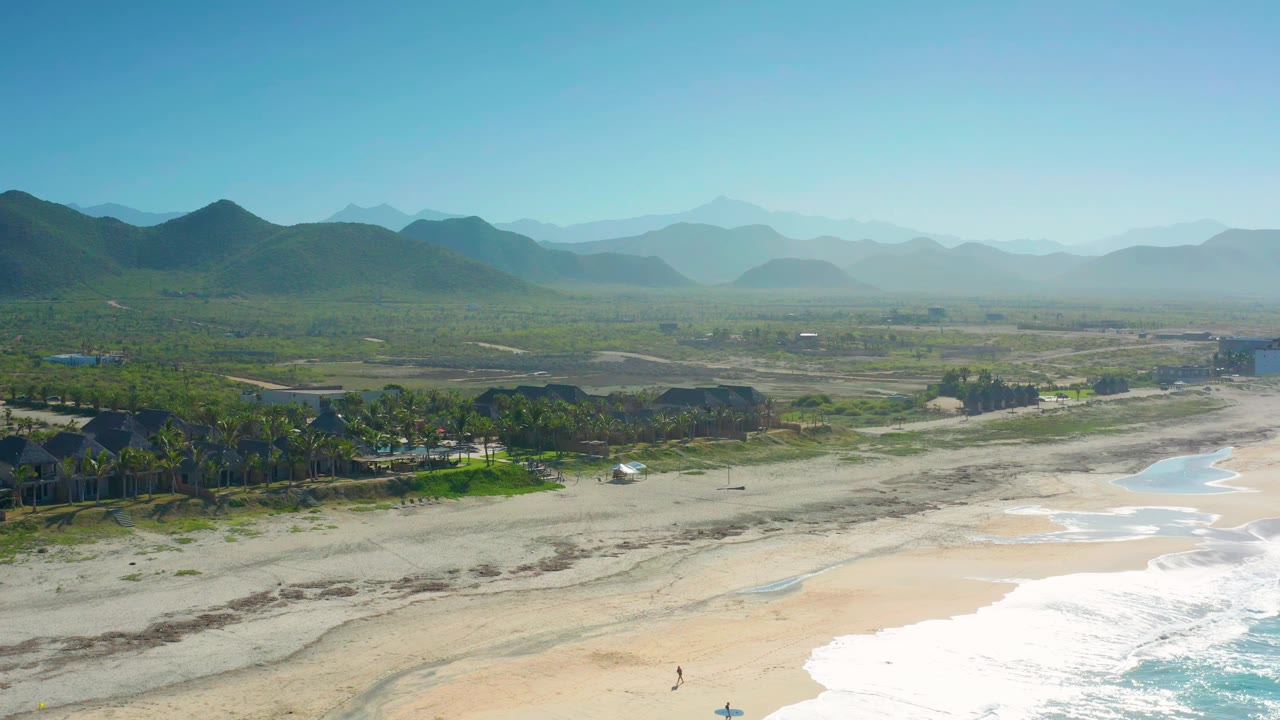 Wide aerial beach road view with surf, palms, buildings, and mountains.