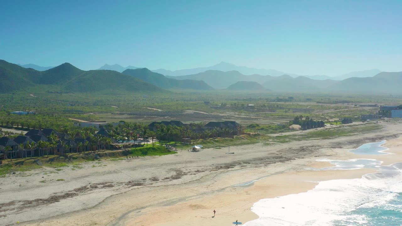 Aerial view in Cerritos with bright surf entering from the lower right, light sand and road tracks across the lower half, a palm-lined built area to the left, layered green hills beyond, and no visible people.