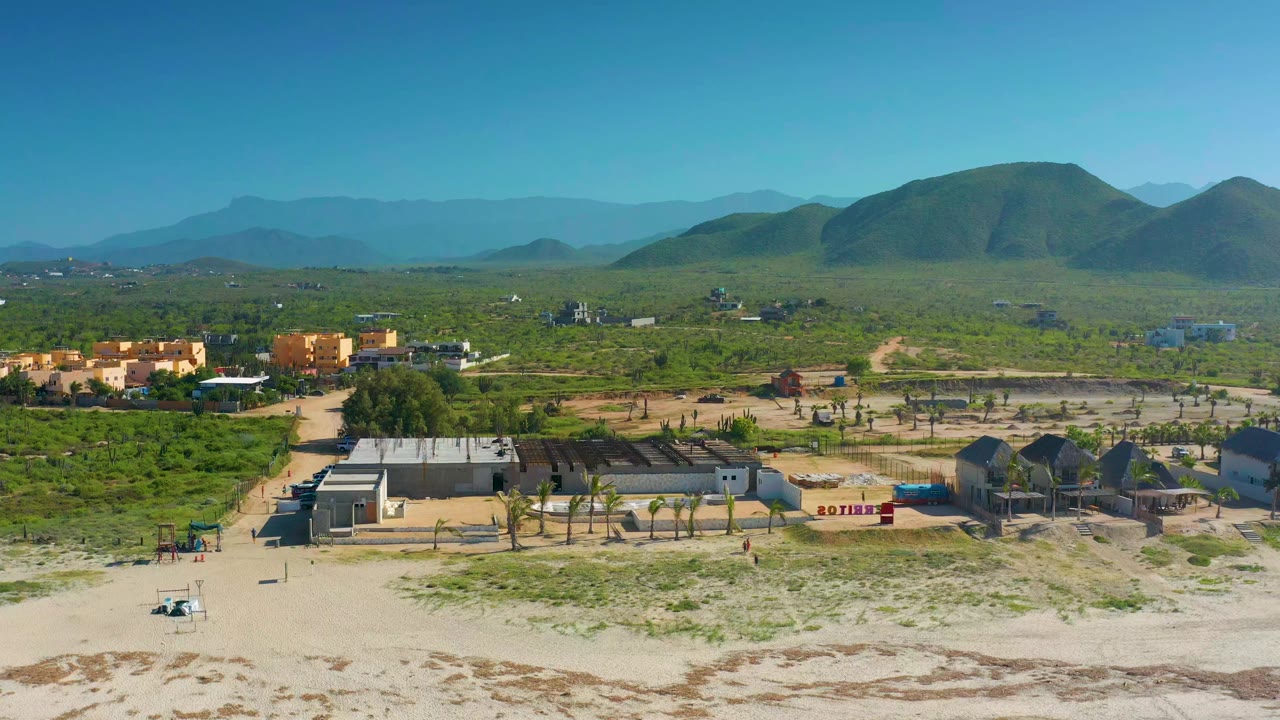 Opening aerial frame in Cerritos, Baja California Sur showing sandy foreground, scattered low houses, scrubland, and rounded green mountains, with no visible people.