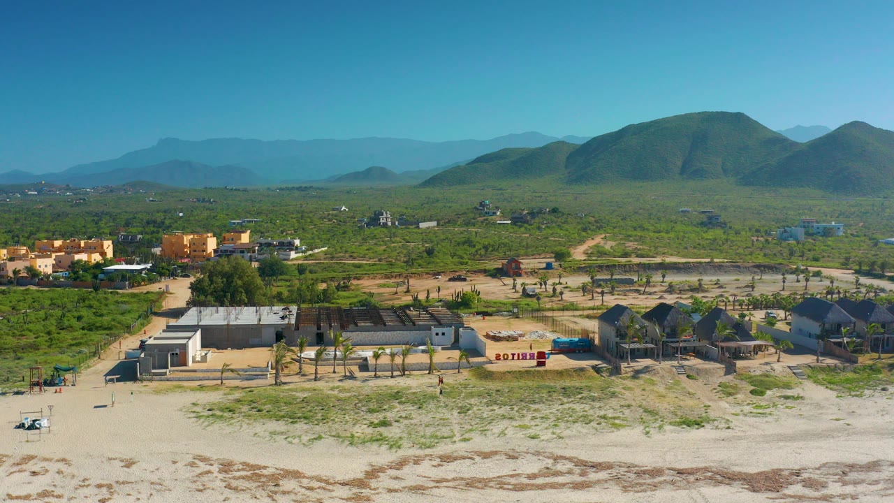 Opening aerial frame over Cerritos, Baja California Sur, showing pale beach sand, low houses, palms, green terrain, rounded mountains, blue sky, and no visible people.