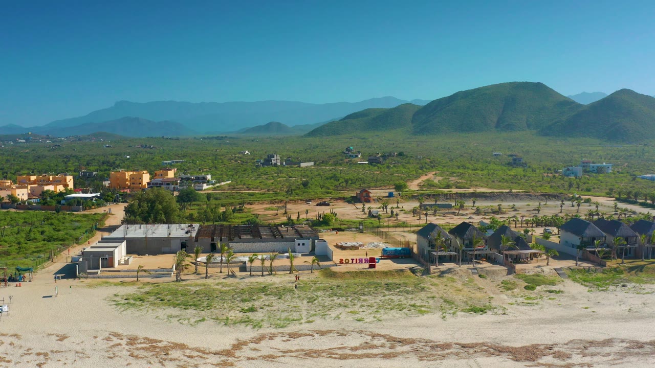 Wide Cerritos aerial with clustered beach houses, palm rows, pale sand at the bottom edge, green vegetation across the plain, mountain forms in back, and no visible people.