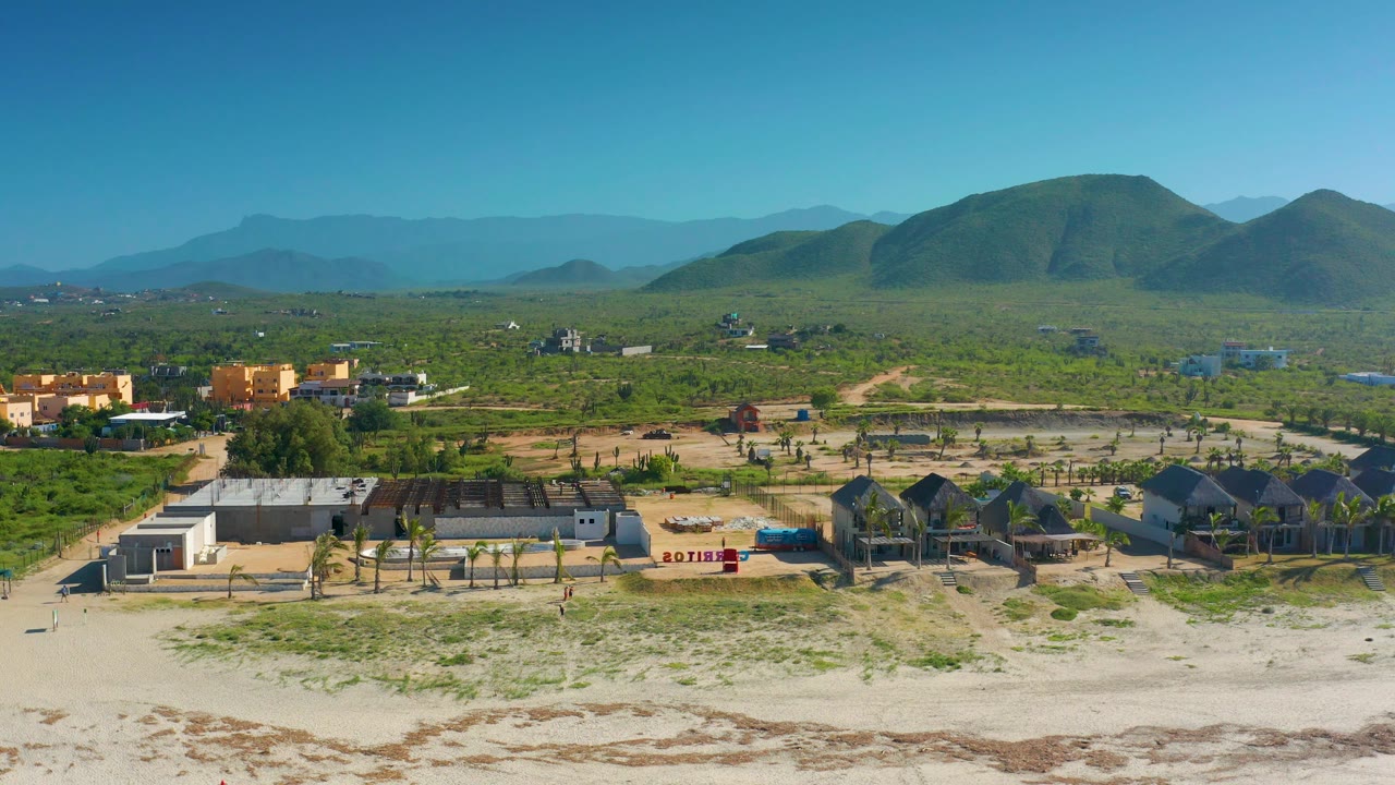 Aerial coastline scene with low buildings, palms, scrubland, and mountains.