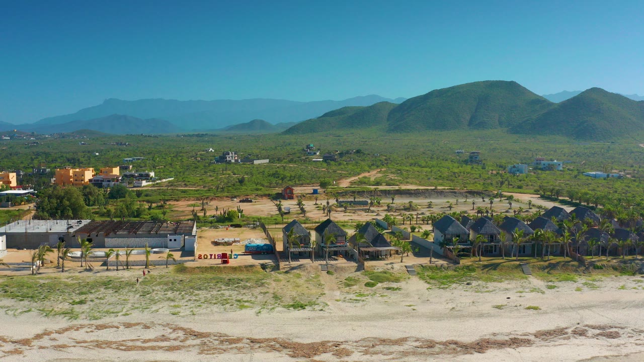 Wide aerial frame over Cerritos, Baja California Sur, Mexico showing pale beach sand, a row of palm huts and low buildings, green scrubland, rounded hills, and no clearly visible people.
