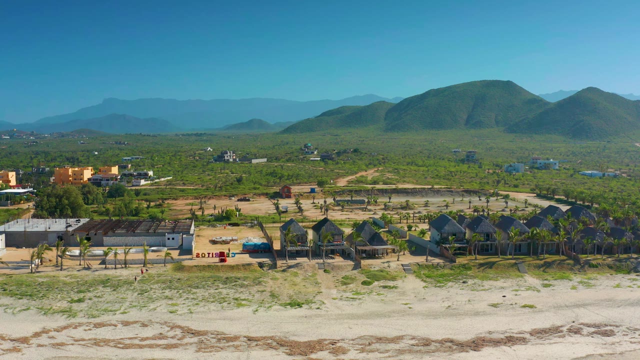 Wide drone view of sandy beach, village structures, palms, and hills.