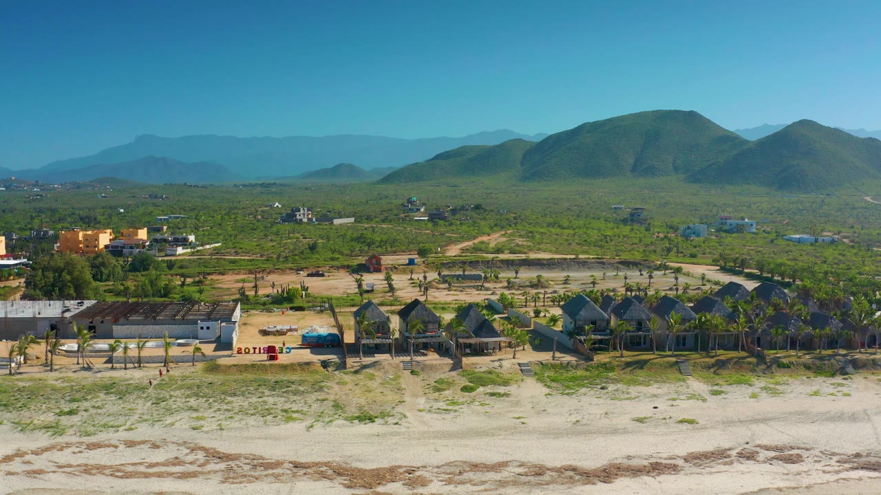 Cerritos aerial still with a pale beach at the bottom, a cluster of huts and rectangular buildings in the midground, and layered green hills fading into blue mountains.