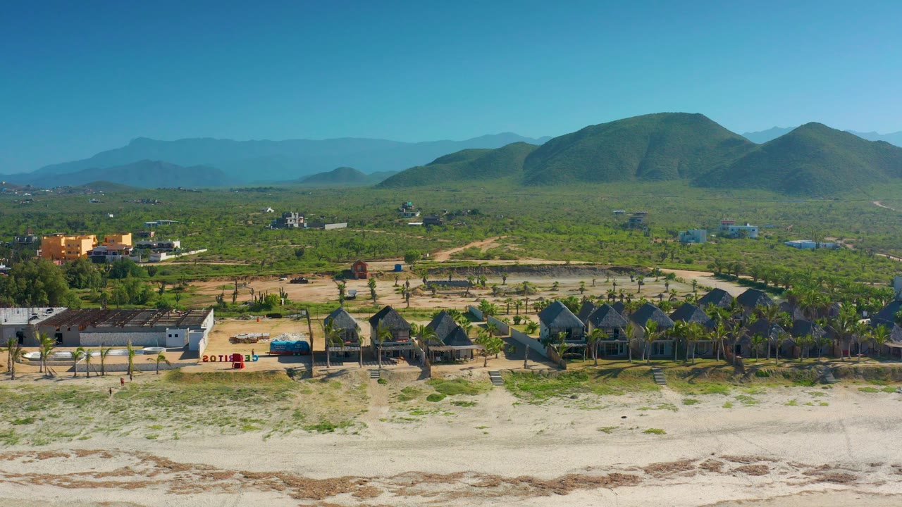 Aerial landscape with beach foreground, palms, low buildings, and hills.