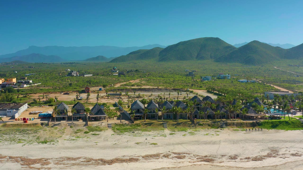 Wide aerial frame in Cerritos, Baja California Sur with beach sand at the bottom, palm-lined holiday buildings near the shore, inland vegetation, distant hills, and no visible people.