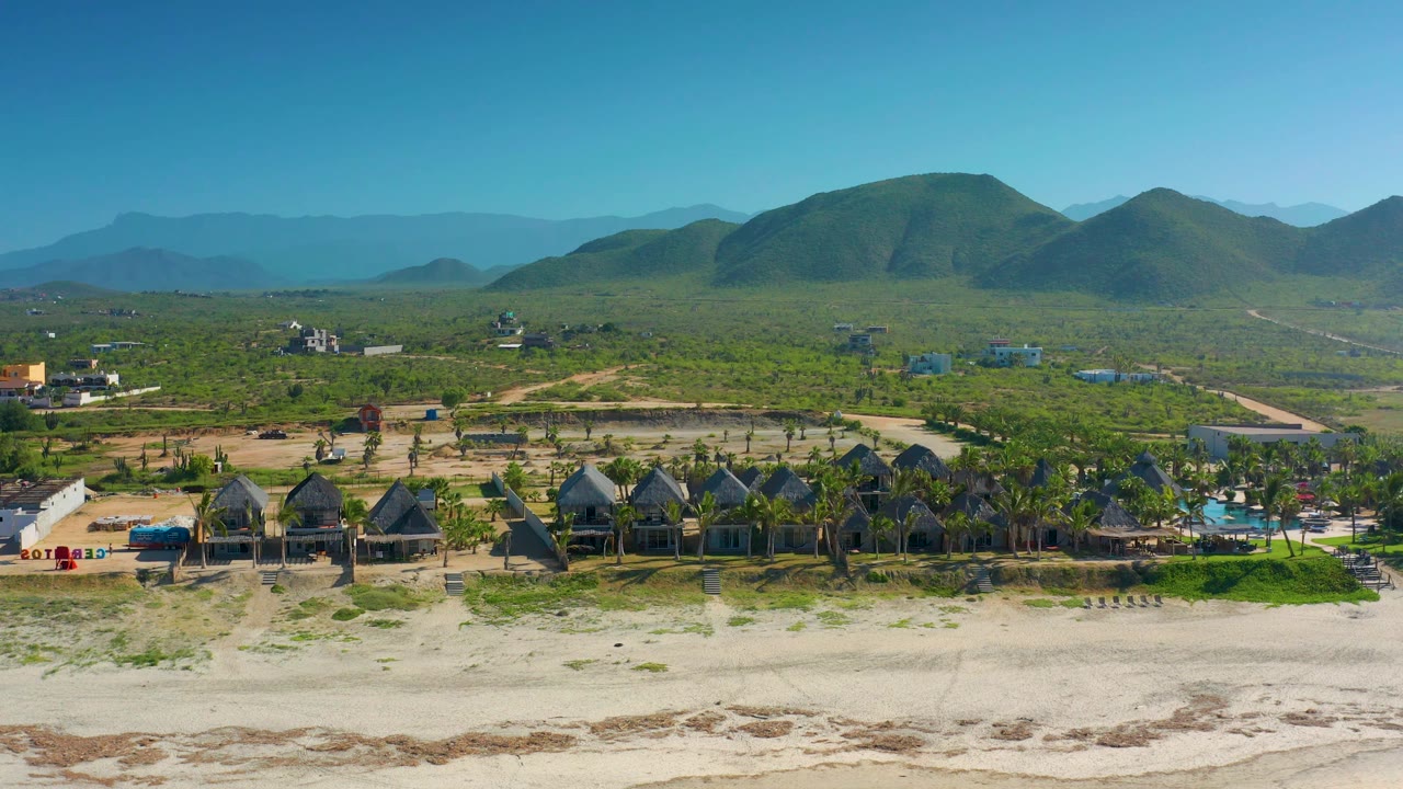 Cerritos, Baja California Sur aerial view with a sandy beach foreground, compact buildings and palms along the shoreline, open green terrain, layered hills, and no visible people.