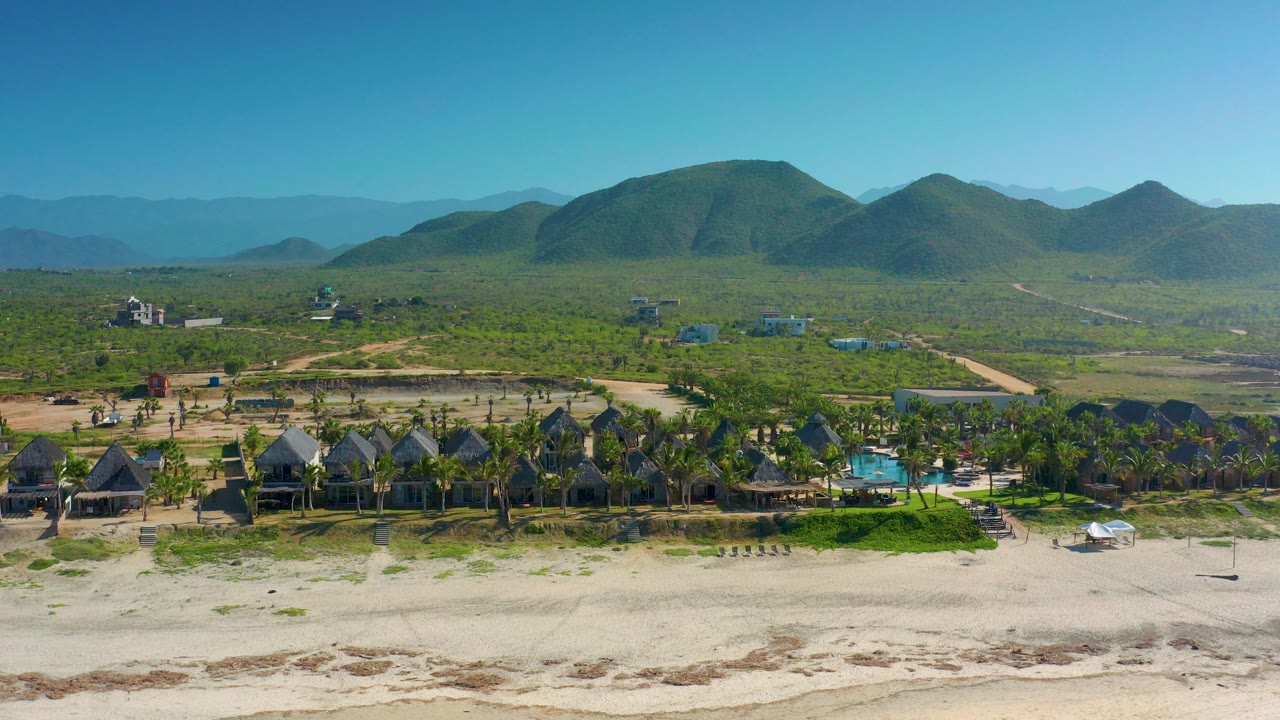 Aerial frame over Cerritos, Baja California Sur showing pale beach sand, hut-like houses, palms, a blue pool, green inland terrain, distant mountains, and tiny people near the beach.