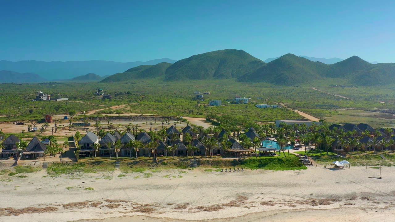 Aerial beach settlement with palms, low houses, pool, and mountains.