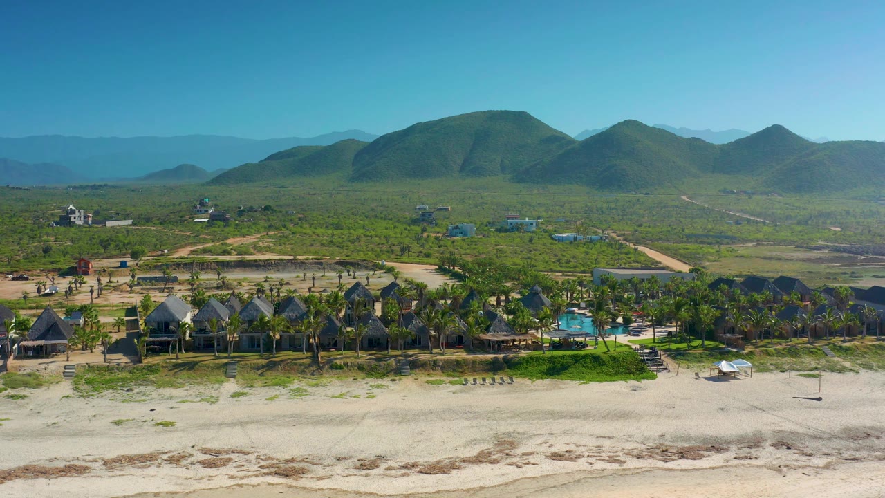 Aerial frame in Cerritos, Baja California Sur with the beach band at the bottom, a cluster of dark-roofed houses and palms, a small blue pool, open green land, and layered hills under a clear sky.