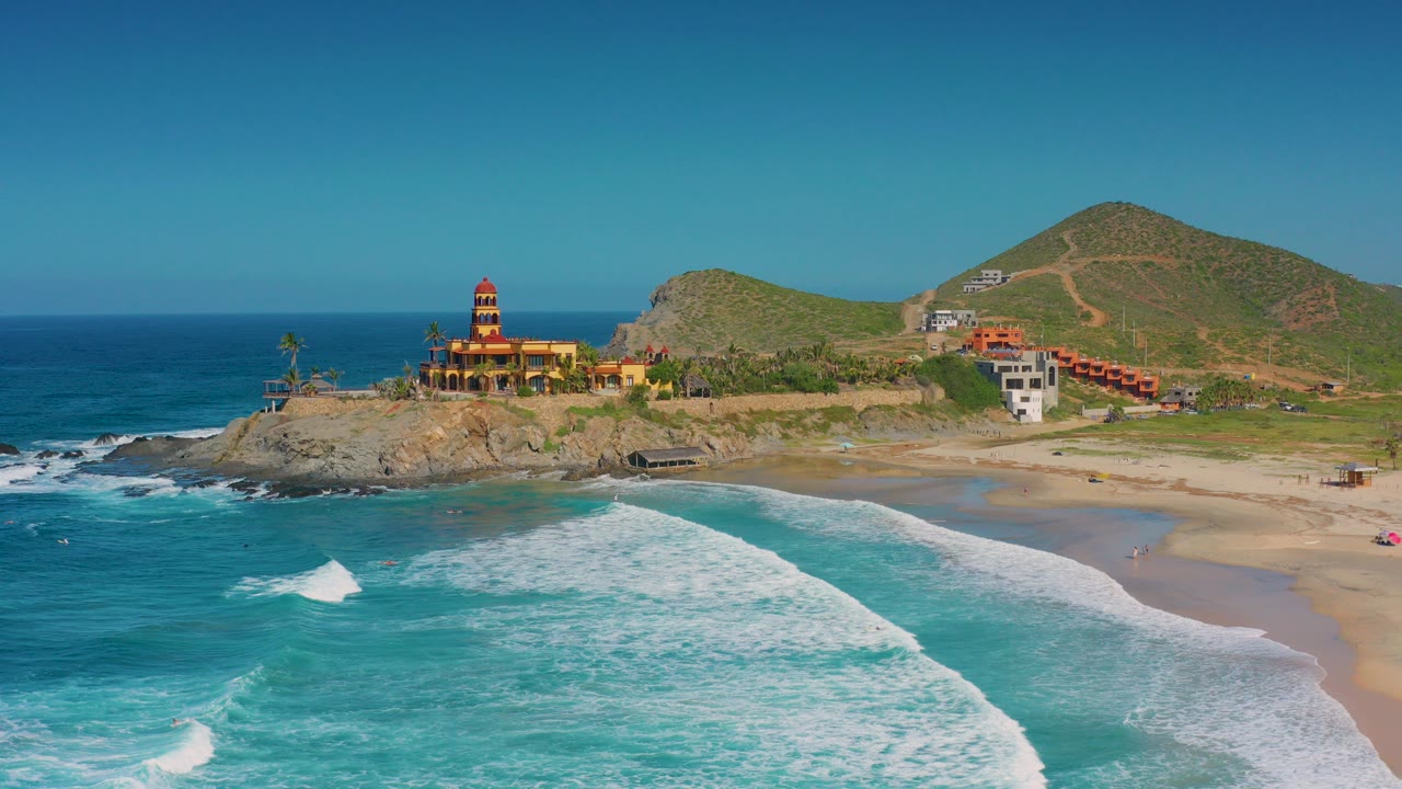 Aerial frame over Cerritos, Baja California Sur, Mexico, showing turquoise waves in the foreground, a sandy beach, a rocky point with coastal buildings, hillside roads, and no visible people.