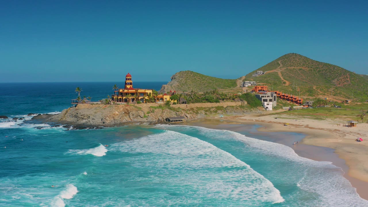 Aerial view in Cerritos with the wave field filling more of the foreground, a sandy cove to the right, the rocky headland and bright tower-like building near the center, and no visible people.