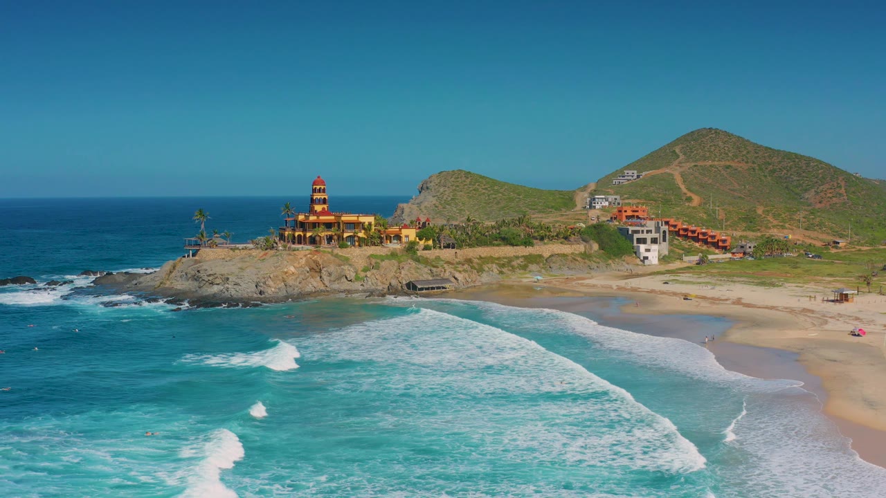 Coastal aerial with turquoise water, white breakers, beach, rocky point, and hillside buildings.