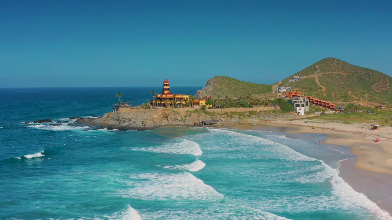 Cerritos aerial frame showing white wave foam spreading across shallow turquoise water, the beach running along the right edge, hillside structures in the distance, and no visible people.
