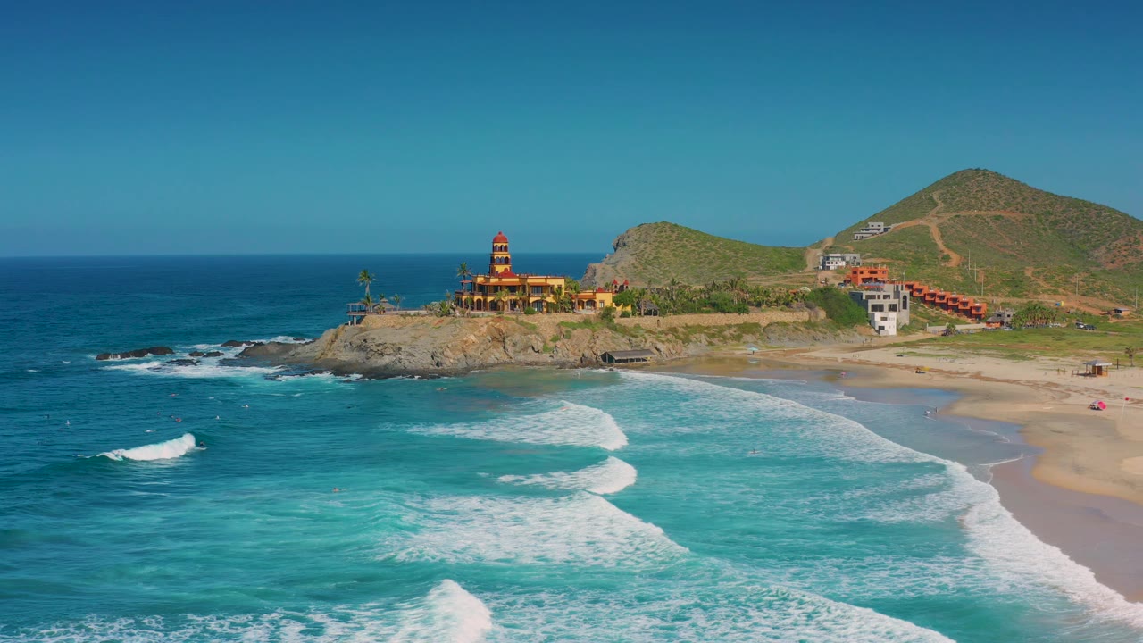 A wide Cerritos coast frame with more open water in the foreground, surf breaking in several white bands, a rocky headland and colorful building near center, hillside roads behind, and no visible people.
