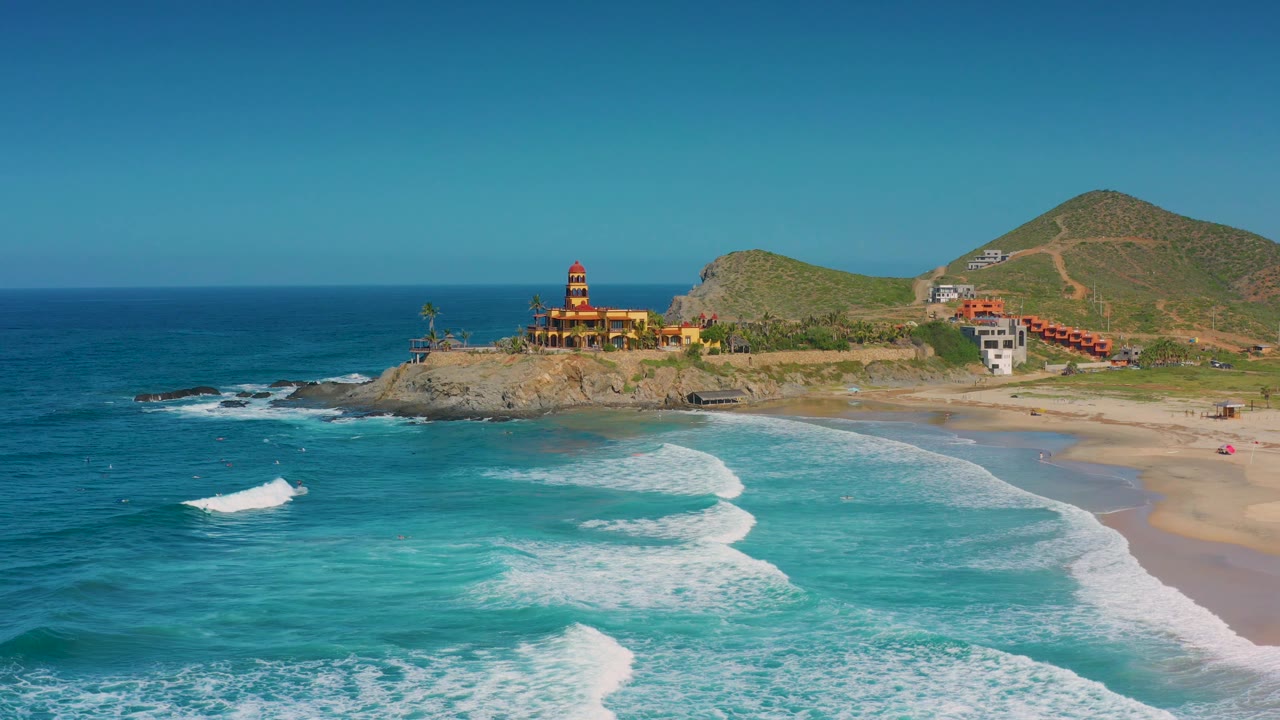 Aerial beach frame with surf in the foreground and coastal buildings near dry hills.