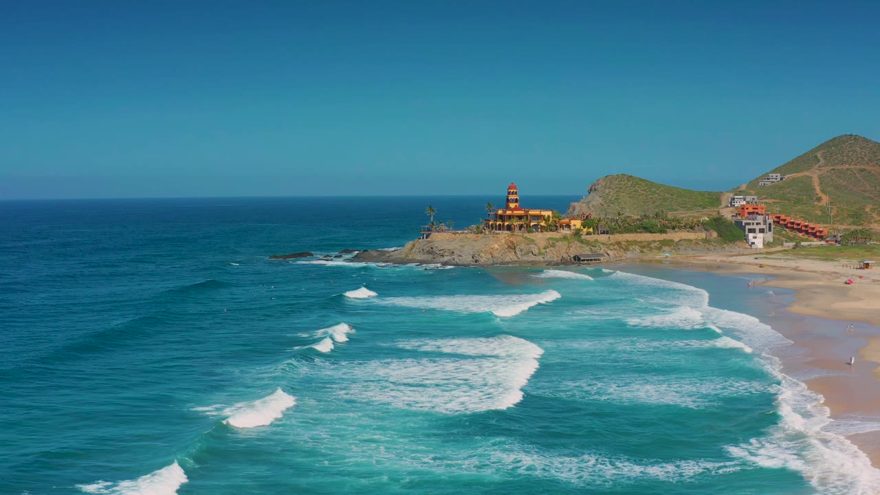 Wide aerial frame of Cerritos coastline showing broad blue water, white breaking waves, sandy beach along the right edge, rocky headland buildings, hillside paths, and no visible people.