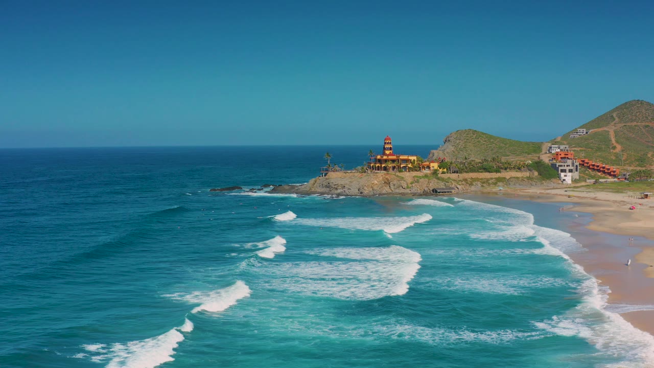 Aerial view in Cerritos, Baja California Sur with diagonal surf lines moving toward the beach, rocky shoreline and coastal buildings near center, hillside terrain behind, and no visible people.
