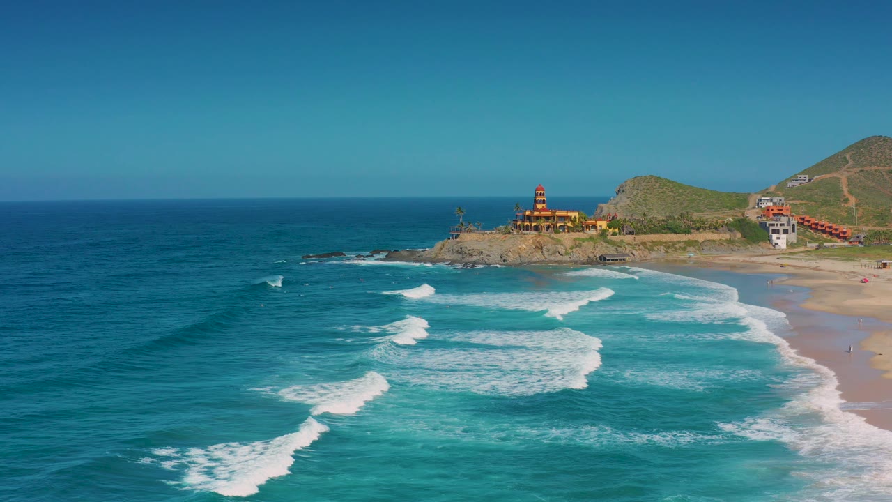 Aerial coastline with large surf patches, sandy beach, rocky point, buildings, and hillside.