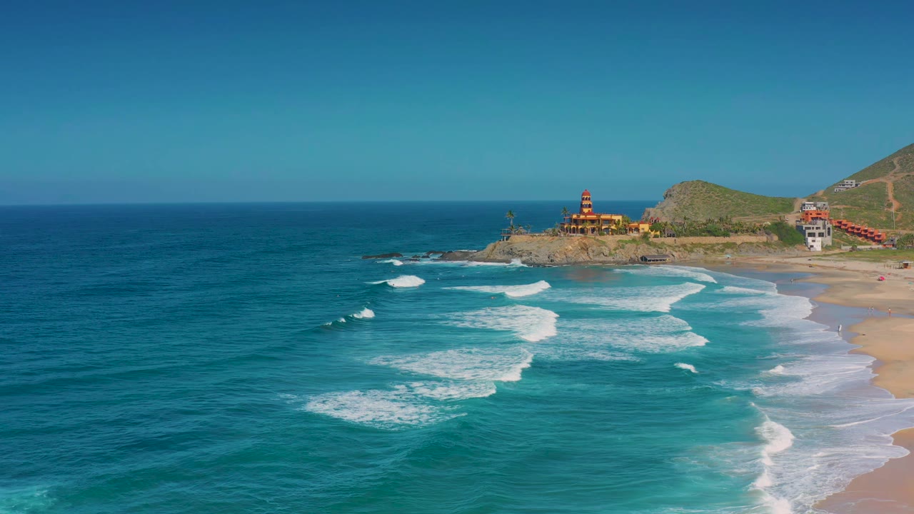 Cerritos coastline seen from above with turquoise shallows spreading toward the beach, larger white wave sets offshore, hillside buildings, and no visible people.